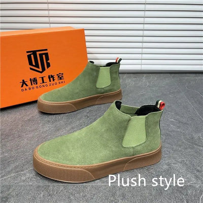 Green Chelsea boots with brown soles in front of an orange box with branding on a gray floor.