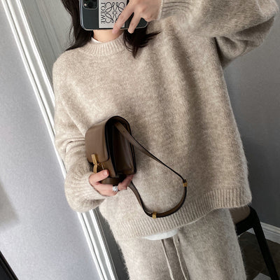 Person wearing a beige sweater holding a brown handbag in front of a mirror.