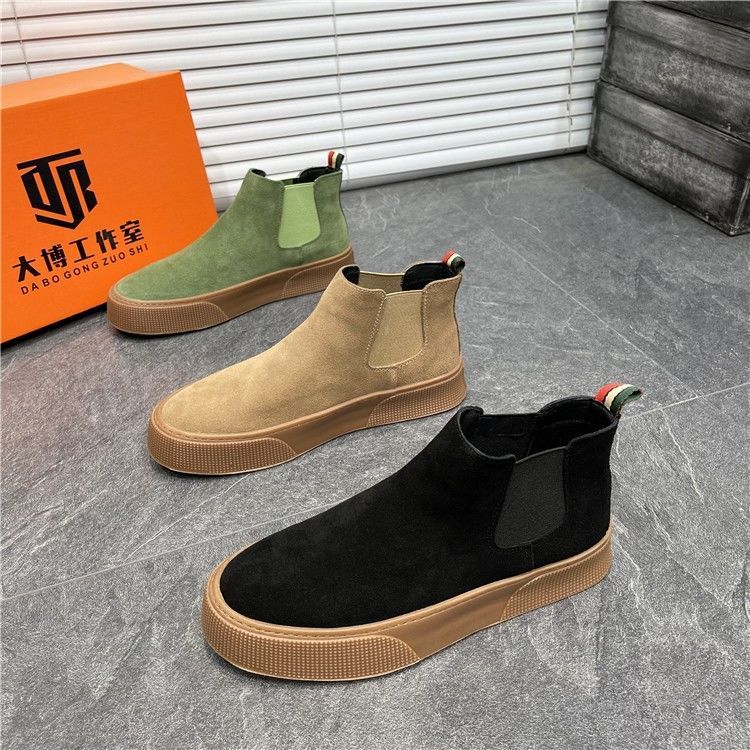 Three suede Chelsea boots in black, brown, and green on a gray floor with an orange box in the background.