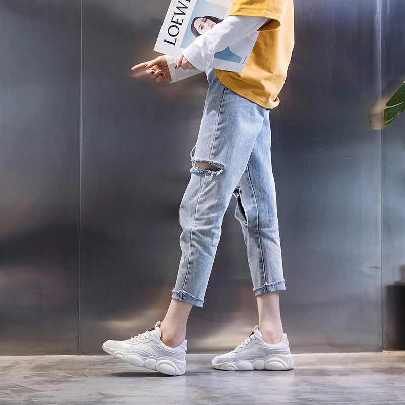 Person wearing light blue ripped jeans and white sneakers, holding a magazine with a blurred background.