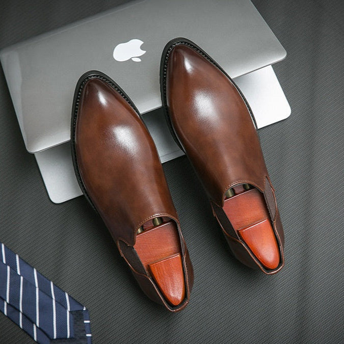 Brown leather shoes with orange insoles on a dark surface with an Apple laptop.