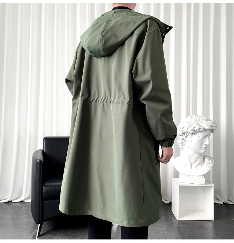 Person wearing a green hooded coat in a room with a black chair and white curtain.