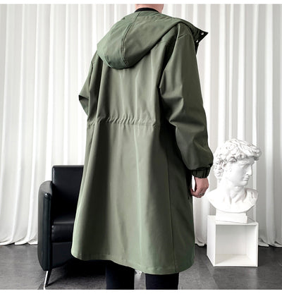 Person wearing a green hooded coat in a room with a black chair and white curtain.