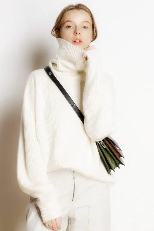 Person wearing a white turtleneck sweater and holding a black handbag against a plain background