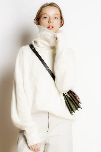 Person wearing a white turtleneck sweater and holding a black handbag against a plain background