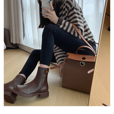 Person wearing brown ankle boots sitting on a wooden floor with a brown handbag.