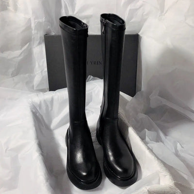 Black knee-high boots on a white textured surface with a brand box in the background.