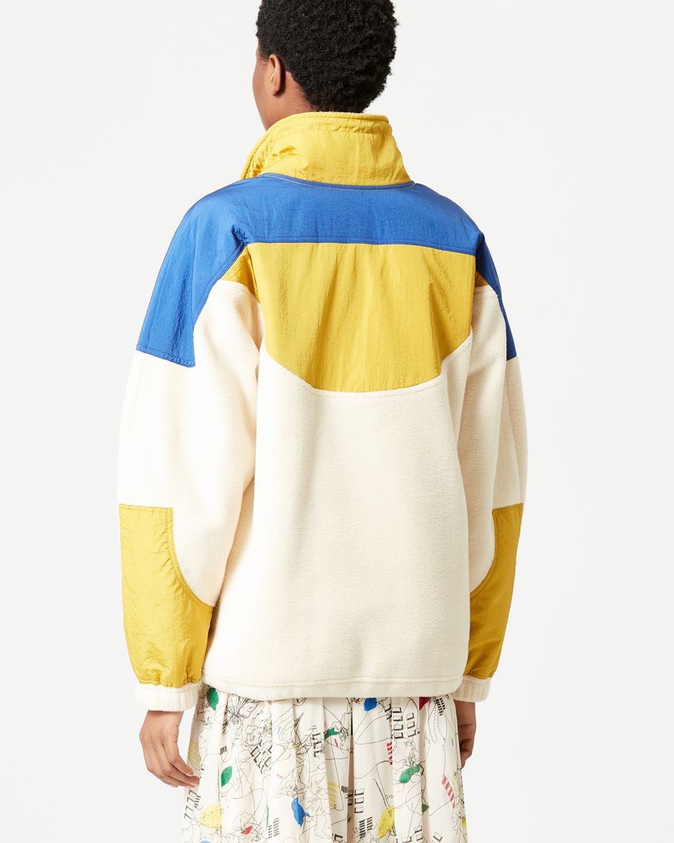 Person wearing a colorful jacket with blue, yellow, and white sections on a plain background