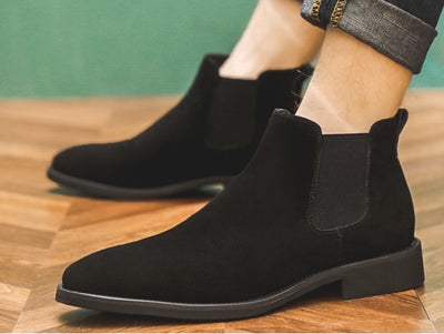 Black suede ankle boots worn on a wooden floor with a green wall background