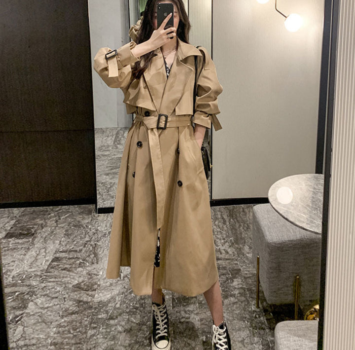 Person wearing a beige trench coat taking a mirror selfie in an indoor setting.