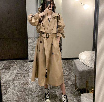 Person wearing a beige trench coat taking a mirror selfie in an indoor setting.