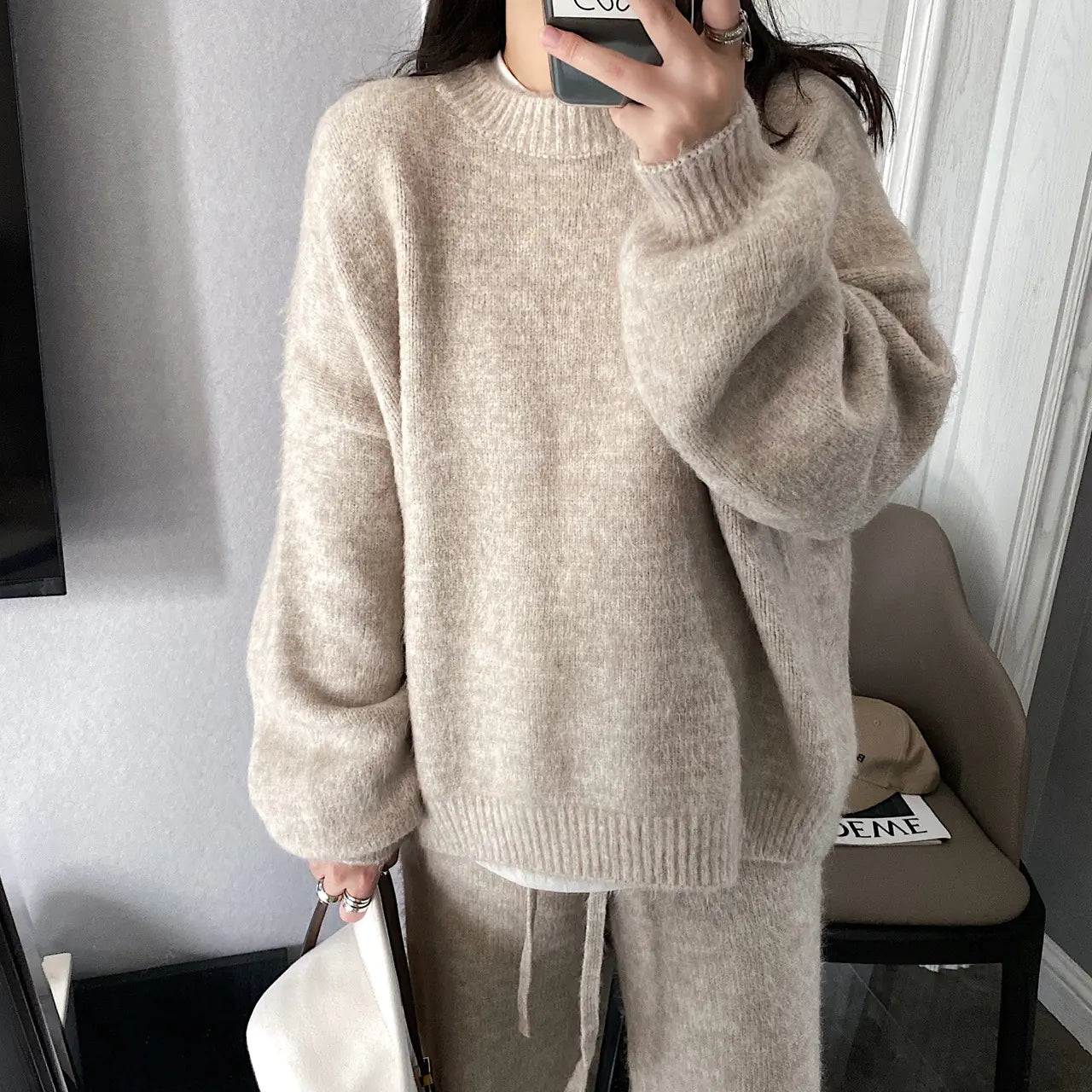 Person wearing a beige sweater and pants in a room with a chair and table.