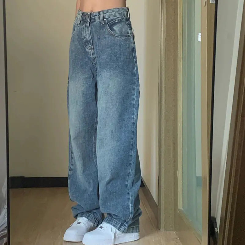 Person wearing blue jeans and white sneakers in a room with a neutral wall and wooden floor.