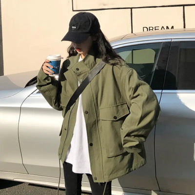 Person wearing a green jacket and black cap standing next to a car, holding a coffee cup.
