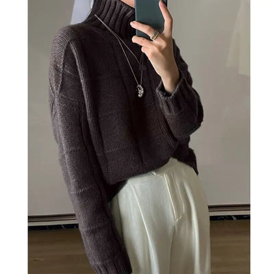 Person wearing a dark brown knitted sweater and light-colored pants, taking a mirror selfie.
