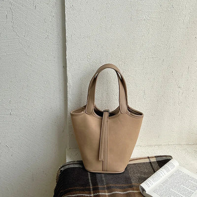 Beige handbag on a textured surface with a plaid fabric and book in the foreground