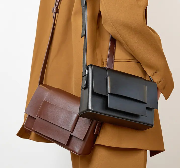 Person wearing a brown coat with a black and brown leather bag over one shoulder on a light background
