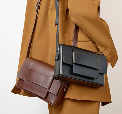Person wearing a brown coat with a black and brown leather bag over one shoulder on a light background