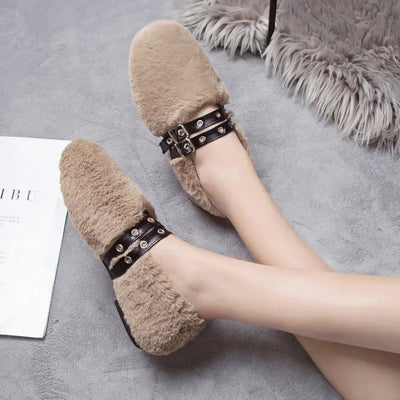 Beige fuzzy slippers with black straps worn by a person on a gray surface.