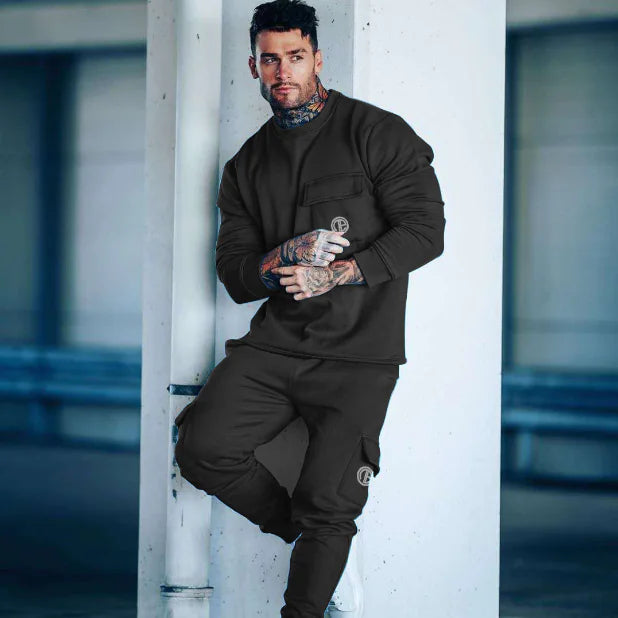Man in black tracksuit leaning against a white column with tattoos visible