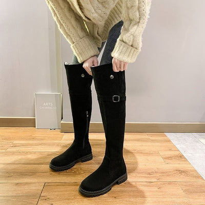 Person wearing black knee-high boots with a beige sweater on a wooden floor.