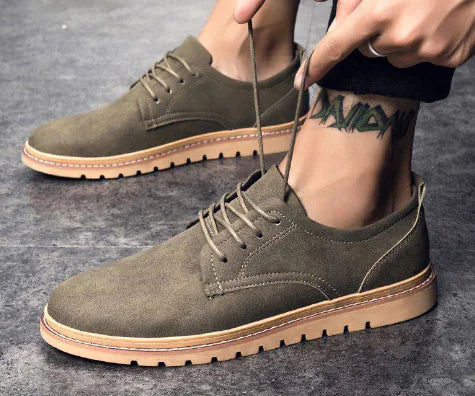 Person wearing olive green suede shoes with brown soles on a gray surface.