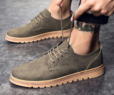 Person wearing olive green suede shoes with brown soles on a gray surface.