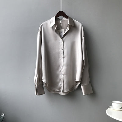 Light gray satin shirt on a hanger against a gray wall with a white table and cup in the corner.