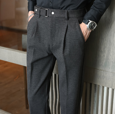 Person wearing a black shirt and gray pleated pants leaning against a wall.
