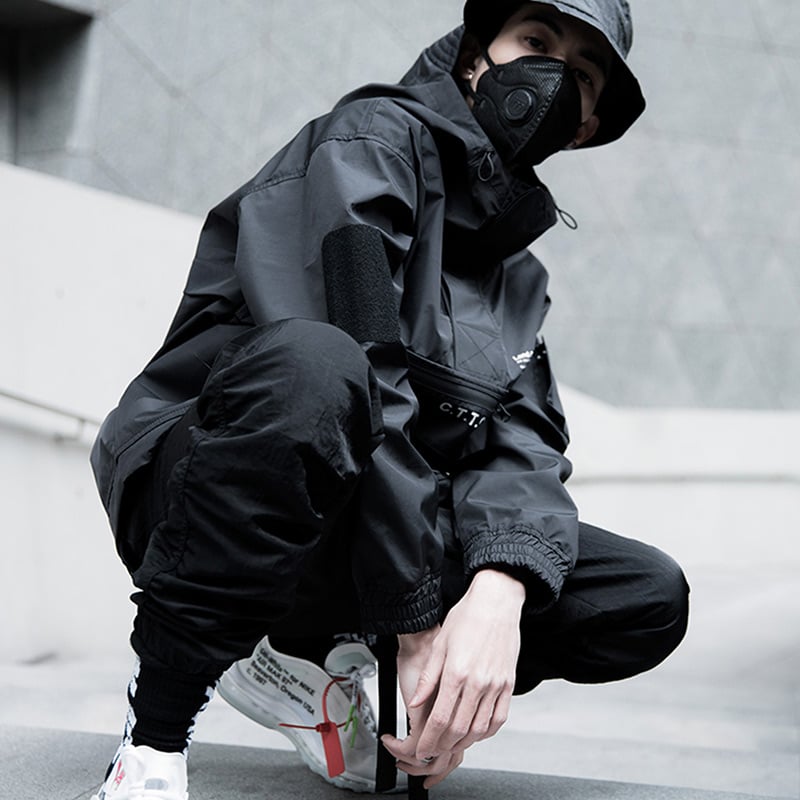 Person wearing a black outfit with a mask, sitting on a concrete surface.