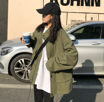 Person wearing a green jacket and black cap holding a coffee cup on a street.