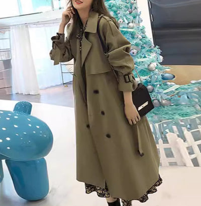 Woman wearing a long beige trench coat in a modern interior setting with a Christmas tree.