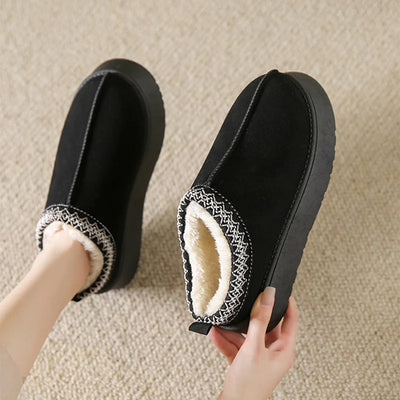 Black slippers with white fur lining held by a person on a beige carpet.