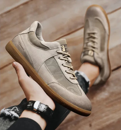 Beige sneakers with brown soles held by a hand on a wooden floor.