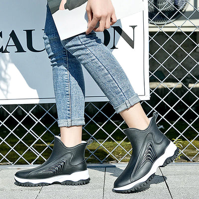 Person wearing black rain boots with white soles on a tiled floor.