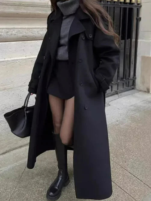 Person wearing a long black coat with a gray sweater and black knee-high boots on a city street.