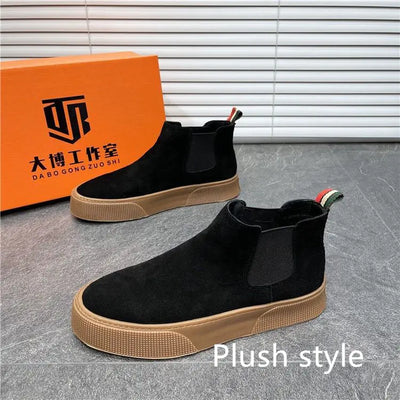 Black Chelsea boots with brown soles next to an orange box on a gray floor.