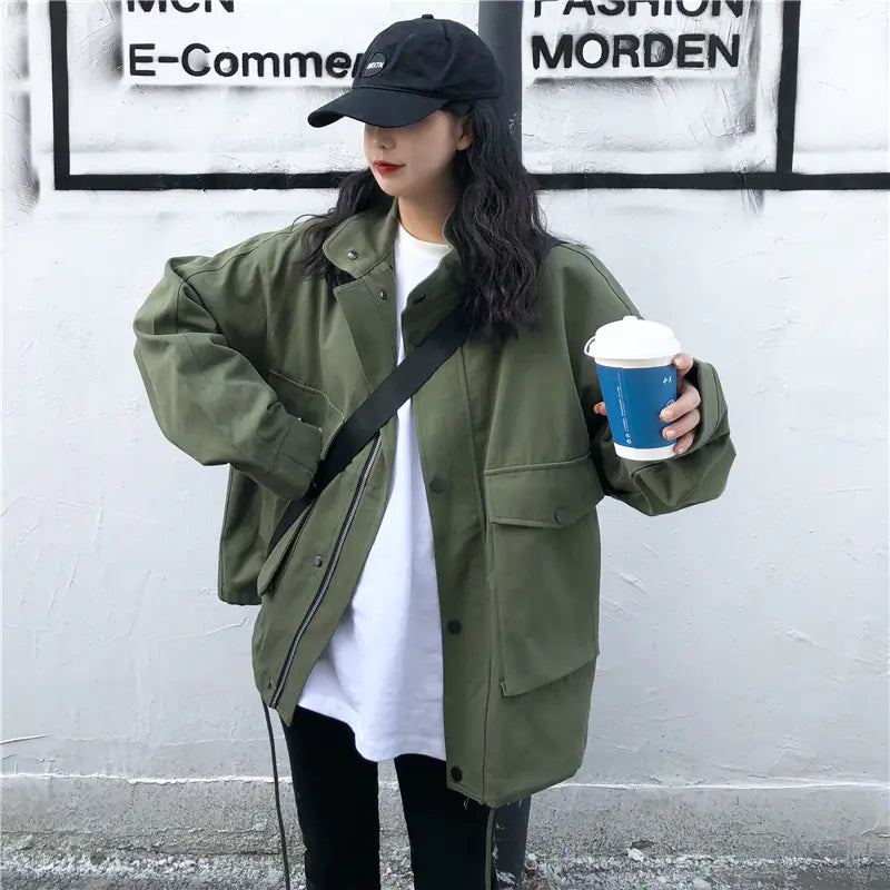 Person wearing a green jacket and black cap, holding a blue cup against a white wall.