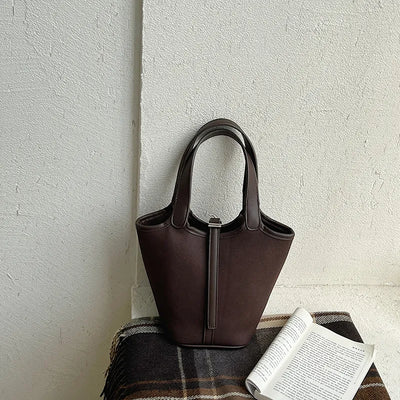 Brown leather handbag on a textured surface with a book and plaid fabric.
